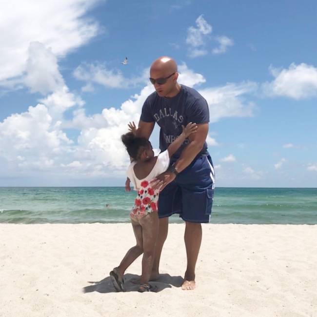 Anthony Ray stands on a sunny beach wearing sunglasses, a navy T-shirt, and shorts, playfully holding a young child as they interact on the sand with the ocean and bright blue sky in the background.
