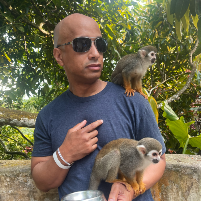 Anthony Ray stands outdoors surrounded by lush greenery, wearing a navy T-shirt and sunglasses, with two small monkeys perched on his shoulder and arm as he looks off to the side with a curious expression.