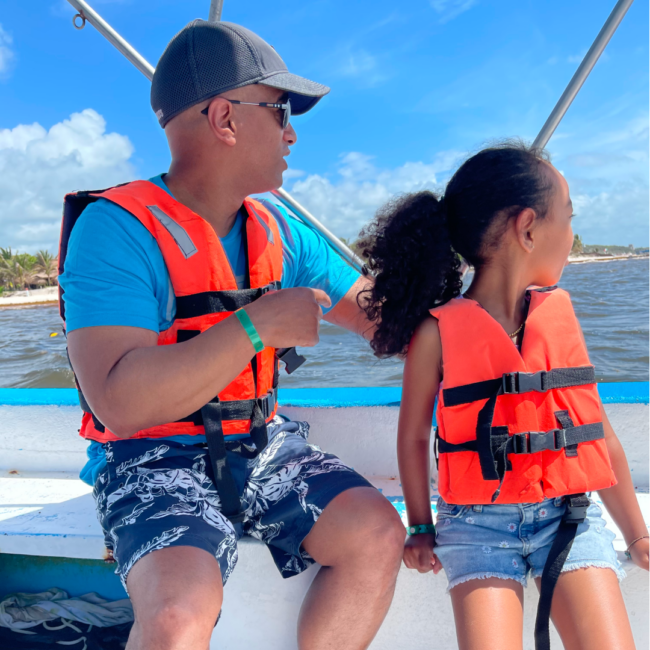 Anthony Ray and a young child sit side by side on a boat, both wearing bright orange life jackets, looking out over the water on a sunny day with a clear blue sky and shoreline in the distance.
