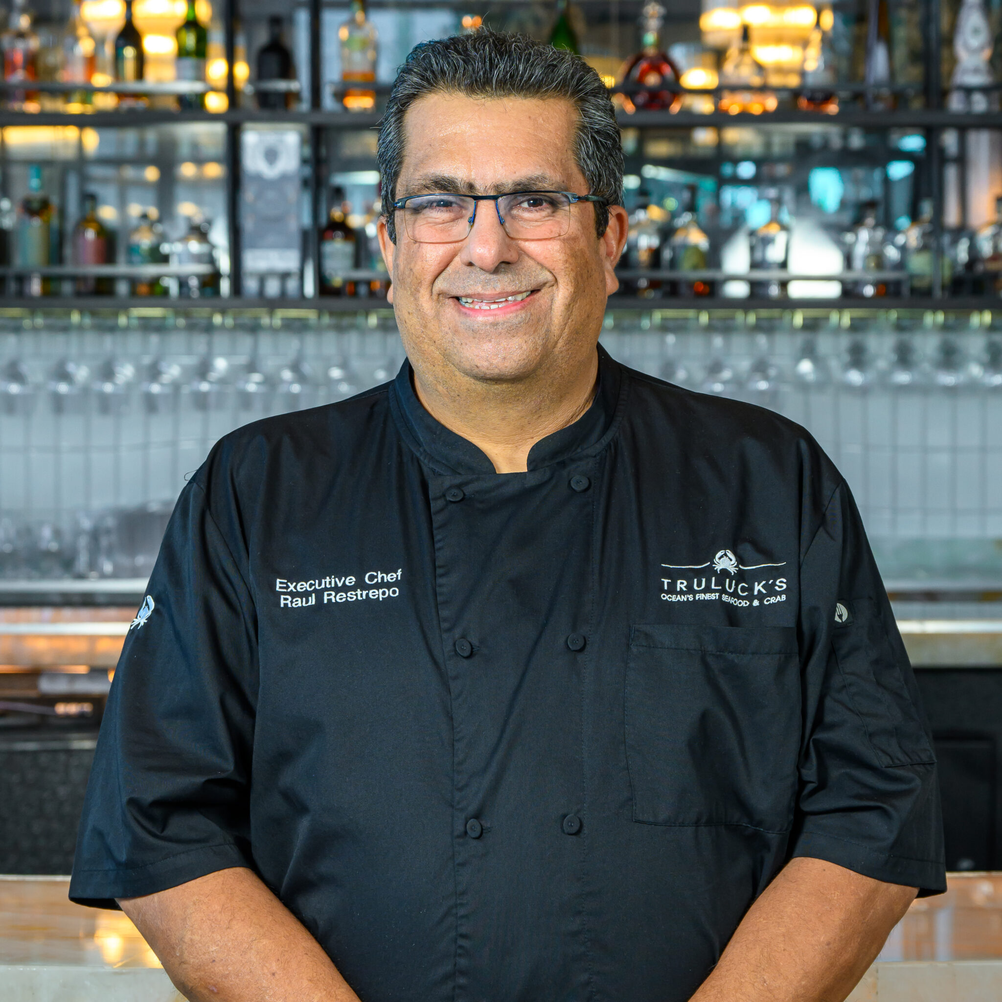 Executive Chef Raul Restrepo of Truluck’s Fort Lauderdale stands smiling in a black chef coat embroidered with his name and the Truluck’s logo, posed in front of a polished bar lined with bottles and glassware.