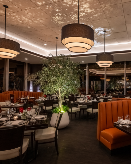 Interior dining room at Truluck’s Miami, featuring elegant table settings, orange banquette seating, warm pendant lighting, and a statement tree centerpiece.