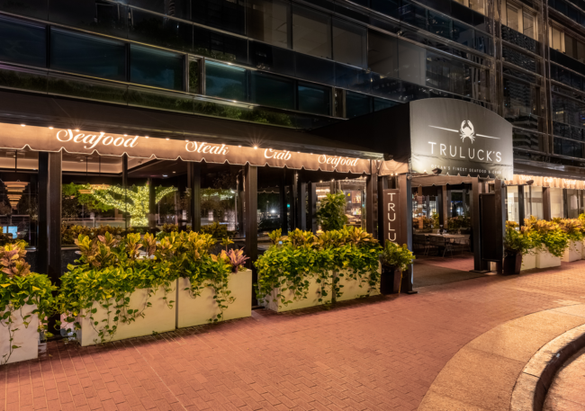 Exterior of Truluck’s Miami at night, featuring illuminated signage, glowing awnings, lush sidewalk planters, and the restaurant’s entrance along the city streetscape.