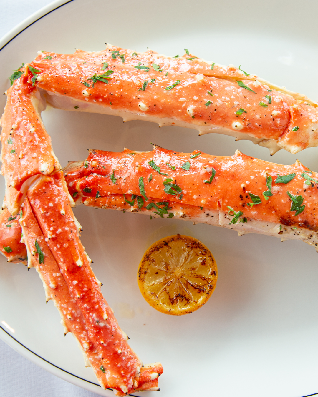 Roasted king crab legs garnished with fresh herbs and served with a charred lemon on a white plate.