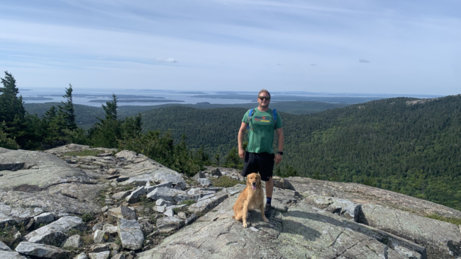 Johnny Griesedieck of Truluck’s Naples standing with a golden retriever on a rocky mountain overlook with forest and water views in the background.