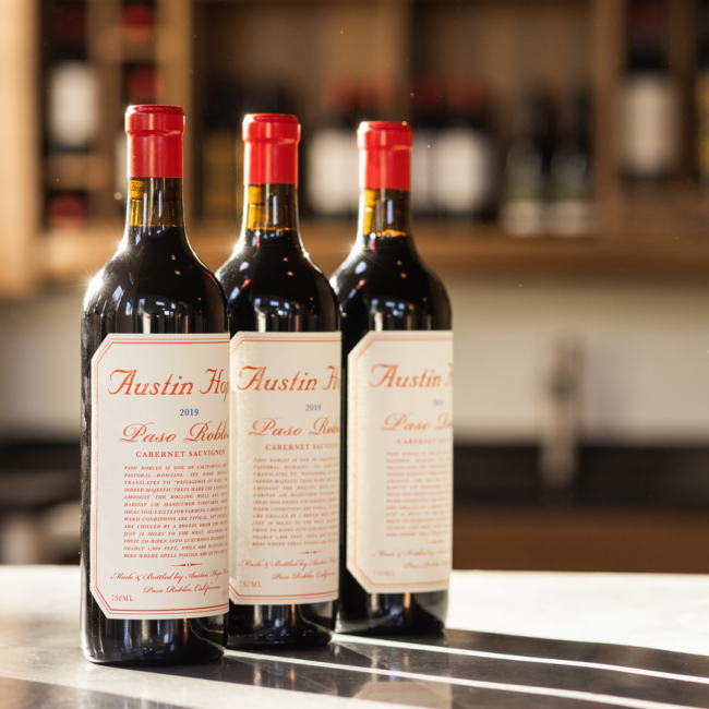 Three bottles of Austin Hope Cabernet Sauvignon are displayed on a bar with a softly blurred wine backdrop.