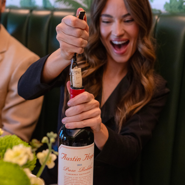 A smiling guest opens a bottle of Austin Hope Cabernet Sauvignon at a restaurant table.