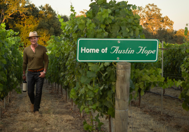 A man wearing a hat walks through a vineyard holding a bottle of Austin Hope wine beside a green “Home of Austin Hope” sign at golden hour.