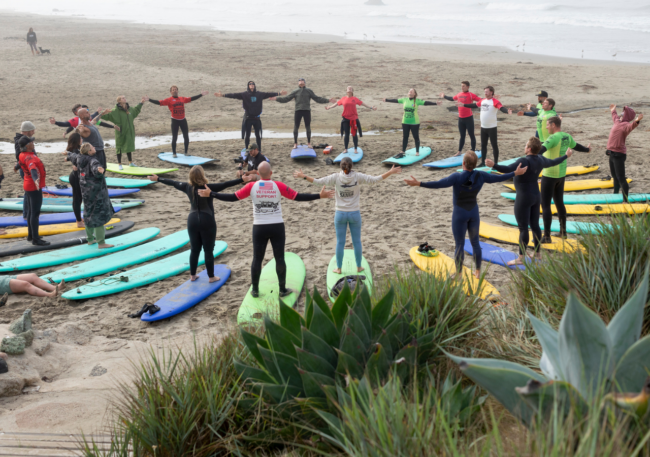 A group of Operation Surf participants and volunteers stand in a circle on a misty beach, arms outstretched in a moment of connection, with colorful surfboards arranged around them on the sand.