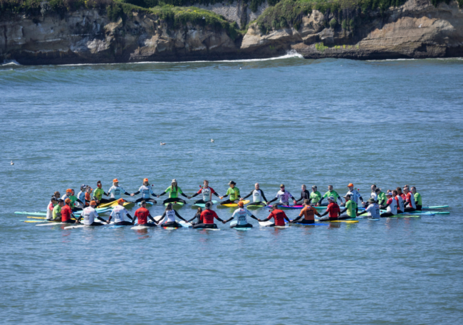 Operation Surf participants and volunteers join hands in a paddle out circle on the ocean, surrounded by surfboards with a rocky coastline in the background.