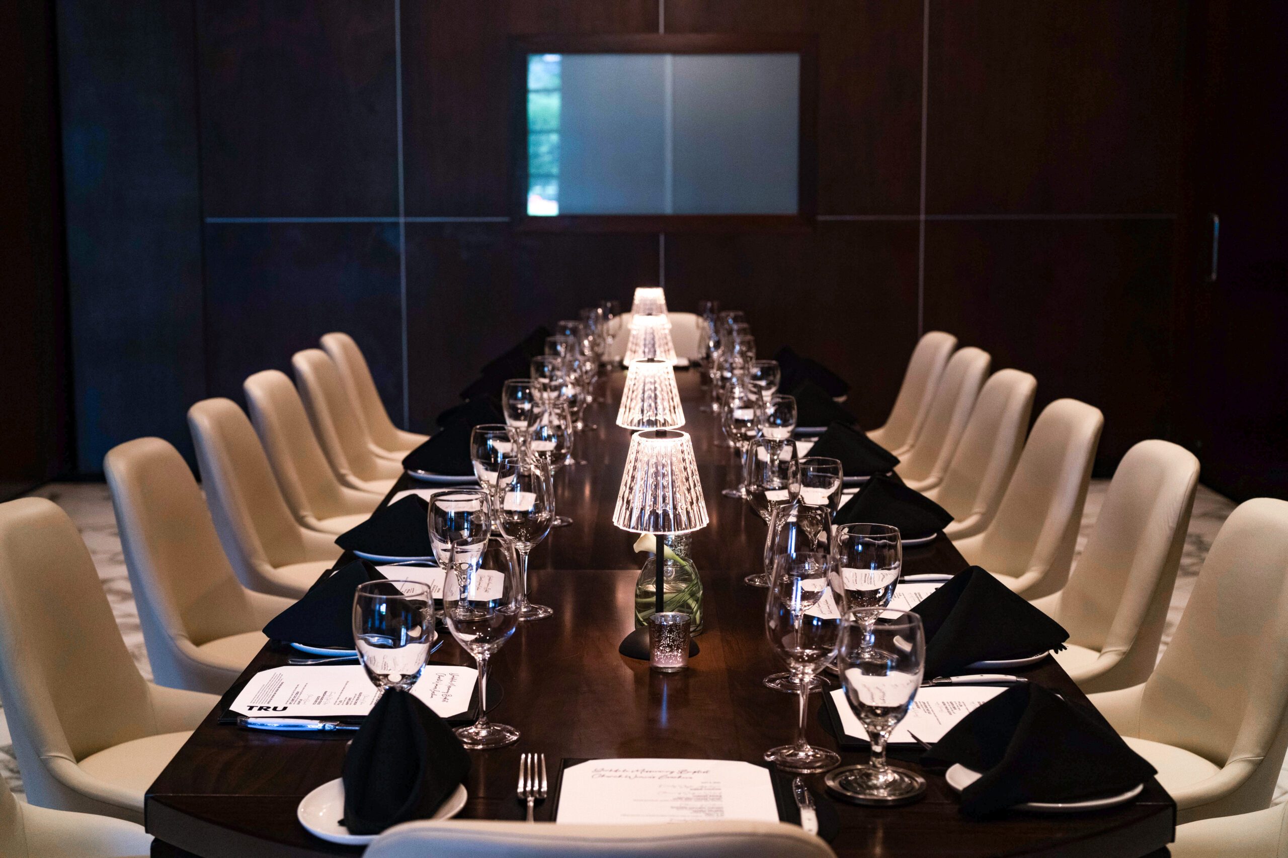 Private dining setup in Truluck’s The Woodlands Capri Room, featuring a long boardroom table with cream chairs, black napkins, glassware, place settings, and softly lit table lamps.