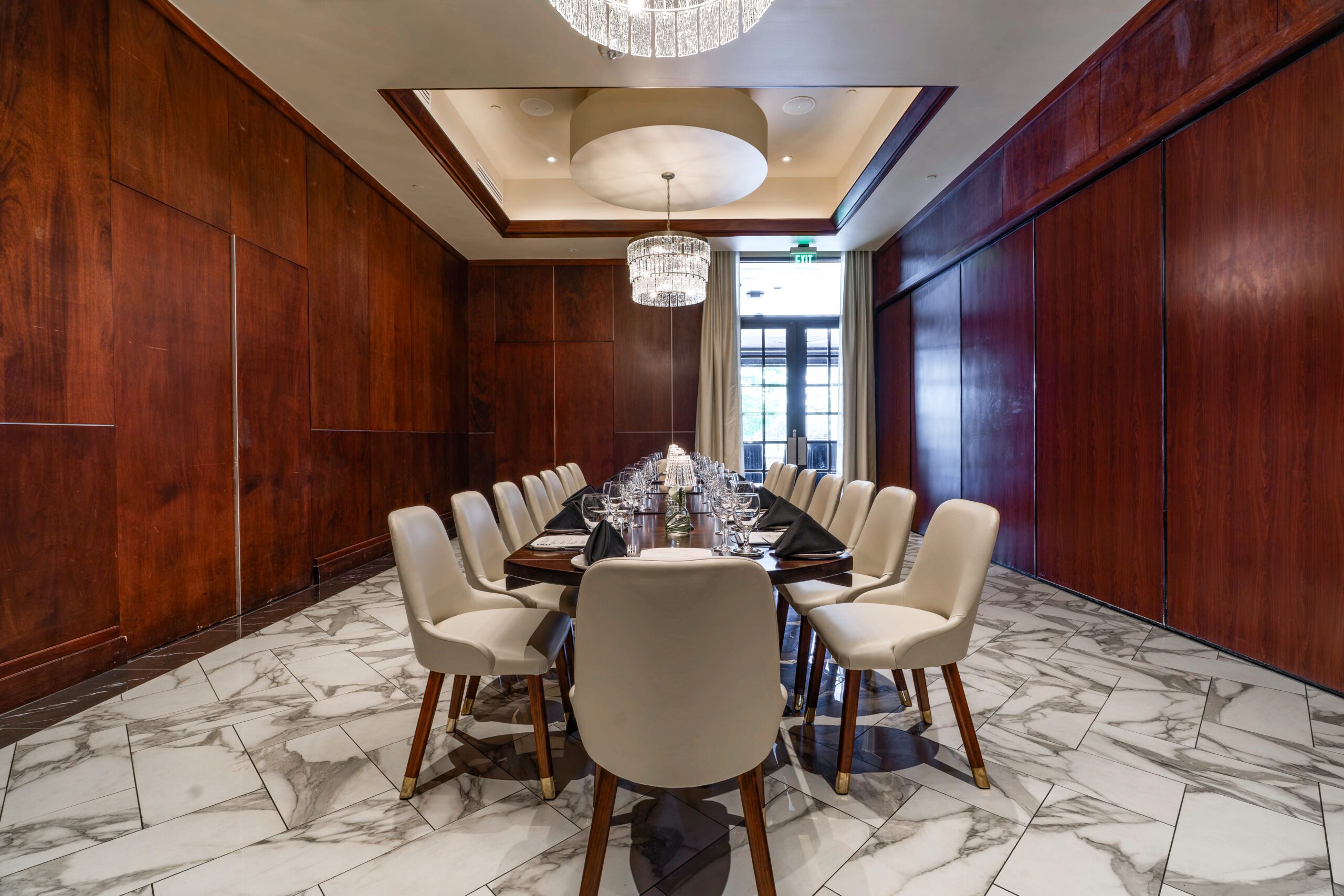 Private dining setup in the Capri Room at Truluck’s The Woodlands, featuring a long boardroom table, cream chairs, rich wood-paneled walls, marble flooring, and elegant chandeliers.