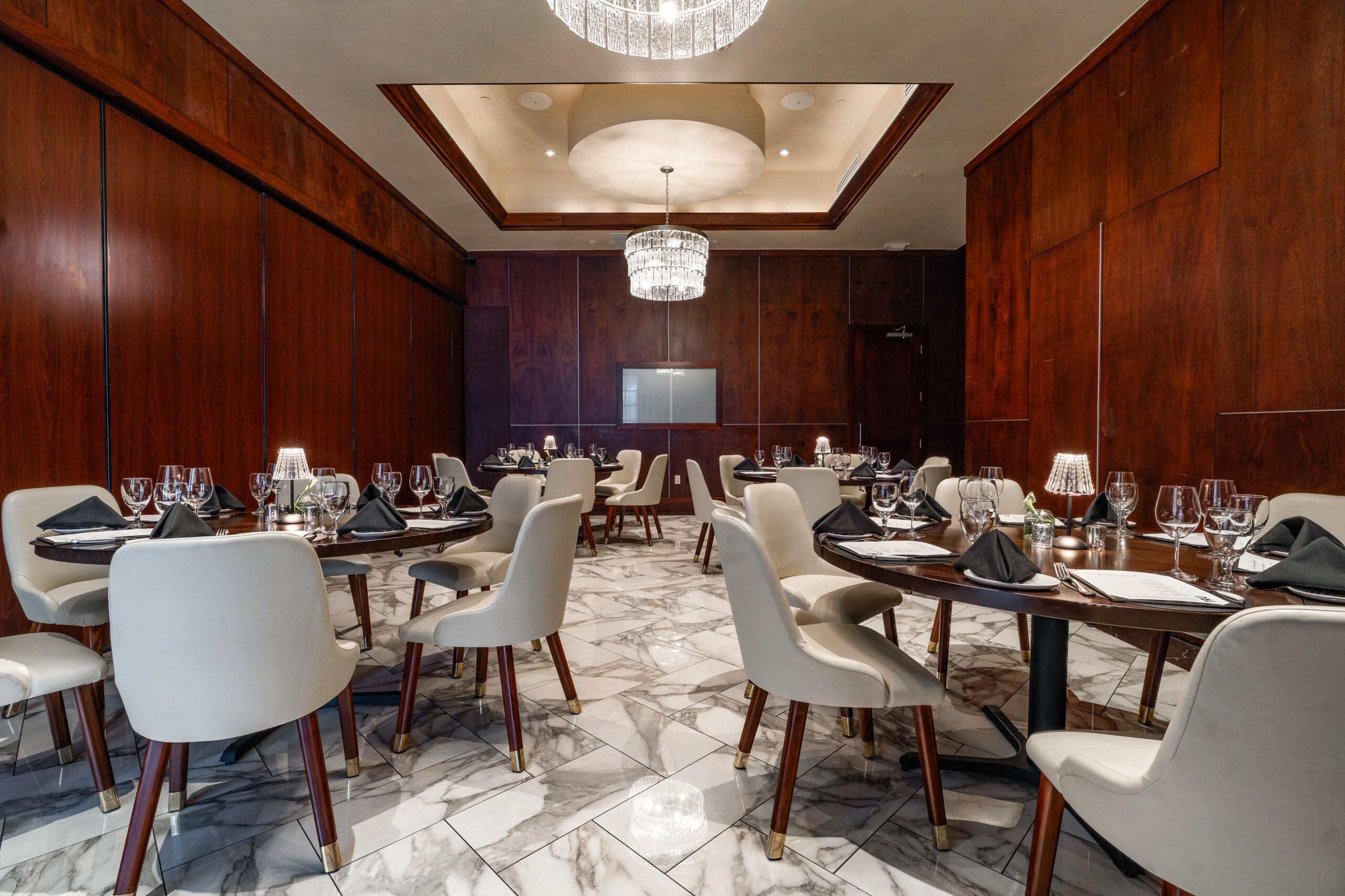 Private dining setup in the Capri Room at Truluck’s The Woodlands, featuring round tables with cream chairs, black napkins, glassware, wood-paneled walls, marble flooring, and crystal chandeliers.