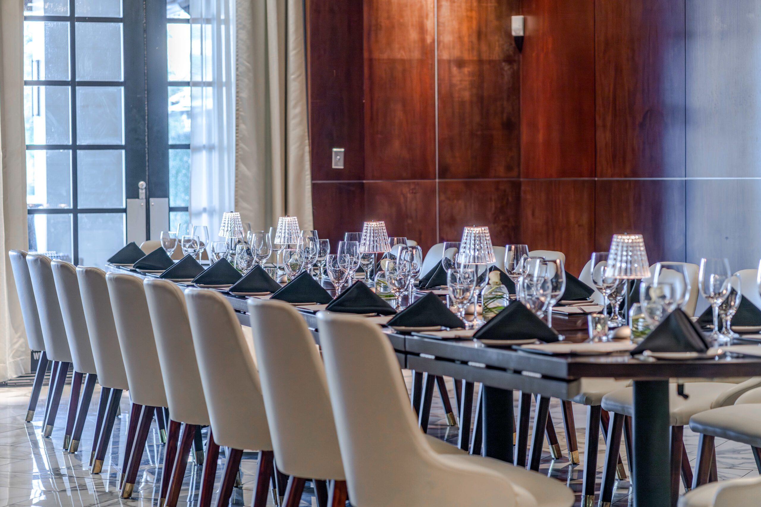Private dining setup in the Naples Room at Truluck’s The Woodlands, featuring a long boardroom table with cream chairs, black napkins, glassware, soft table lamps, wood-paneled walls, and floor-to-ceiling windows.