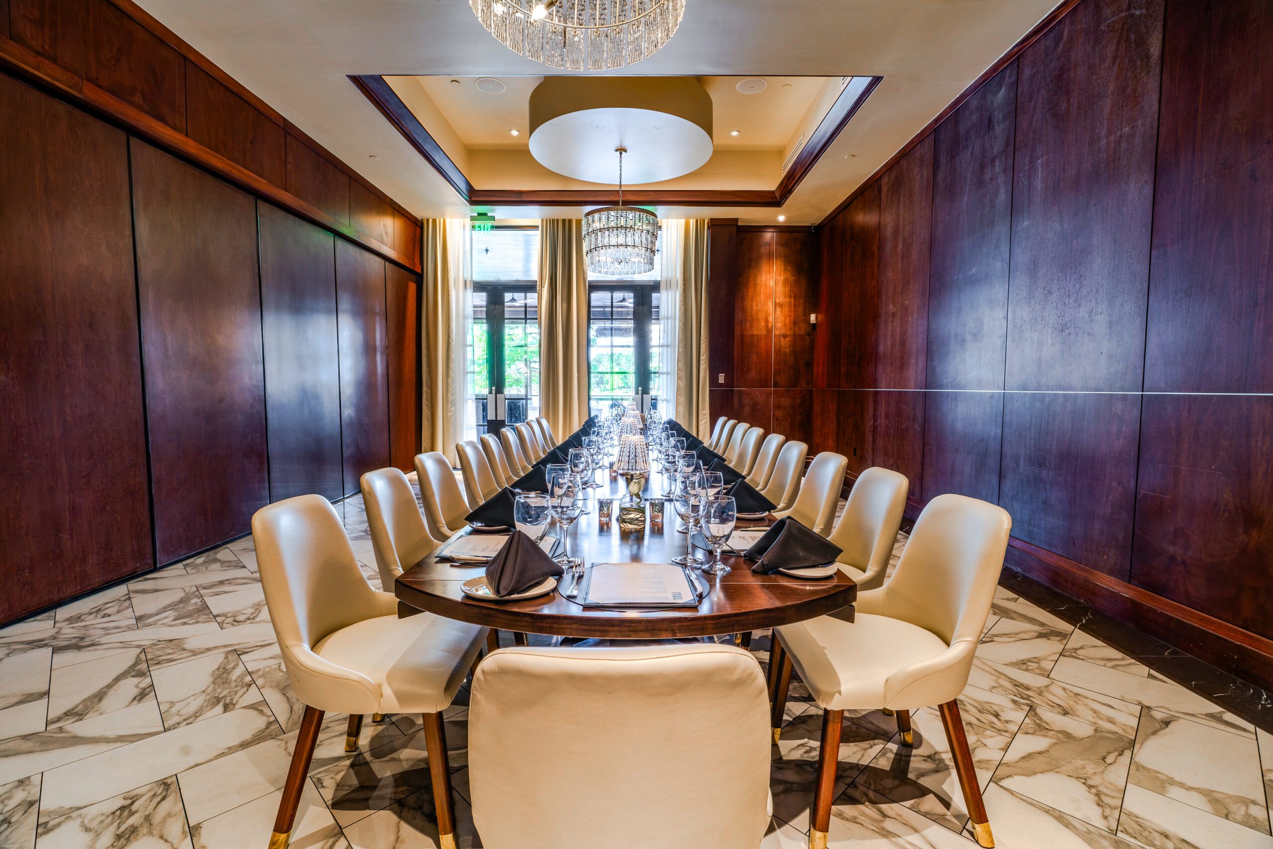 Private dining setup in the Naples Room at Truluck’s The Woodlands, featuring a long boardroom table with cream chairs, black napkins, glassware, wood-paneled walls, marble flooring, and crystal chandeliers.