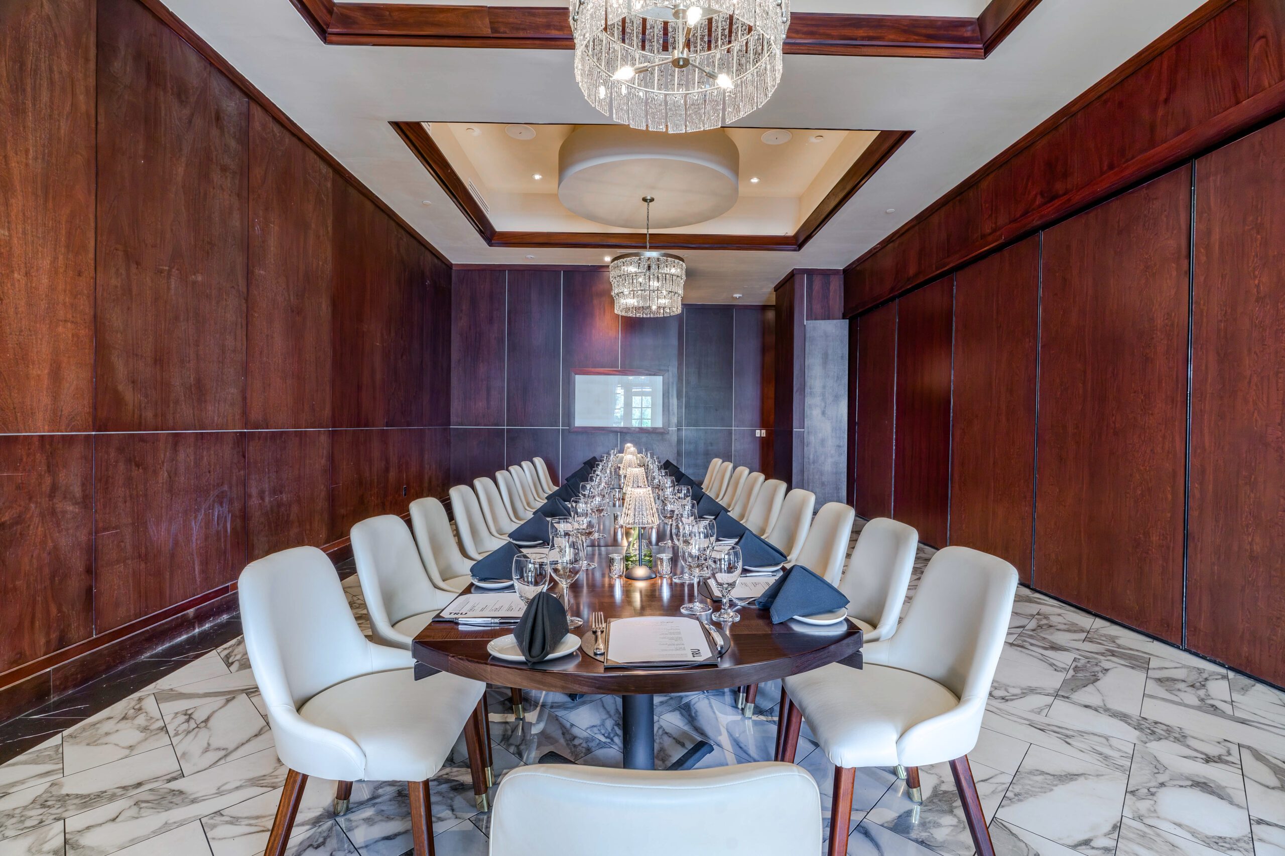 Private dining setup in the Naples Room at Truluck’s The Woodlands, featuring a long boardroom table with cream chairs, black napkins, glassware, rich wood-paneled walls, marble flooring, and crystal chandeliers.