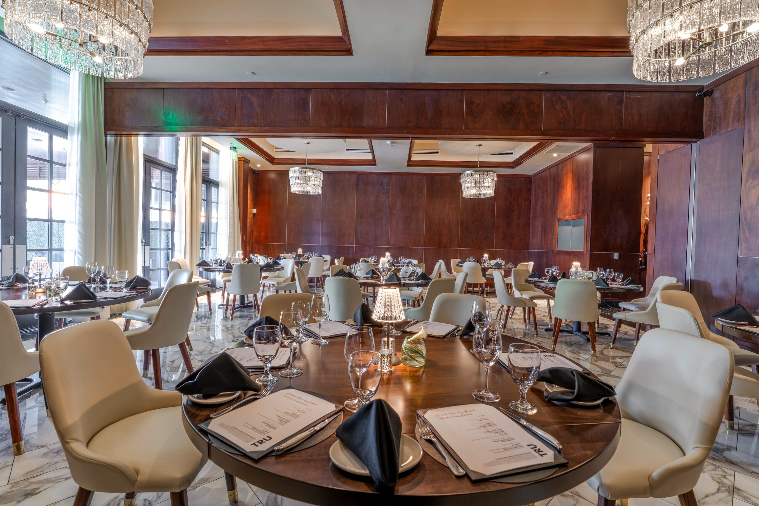 Private dining setup in the Stone Crab Room at Truluck’s The Woodlands, featuring round tables with cream chairs, black napkins, glassware, soft table lamps, wood-paneled walls, marble flooring, and crystal chandeliers.