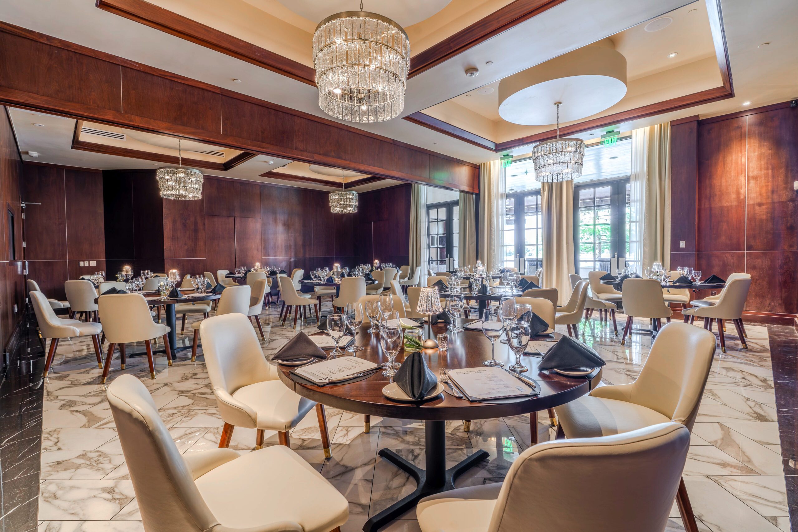 Private dining setup in the Naples Room at Truluck’s The Woodlands, featuring round tables with cream chairs, black napkins, glassware, crystal chandeliers, wood-paneled walls, marble flooring, and natural light from tall windows.