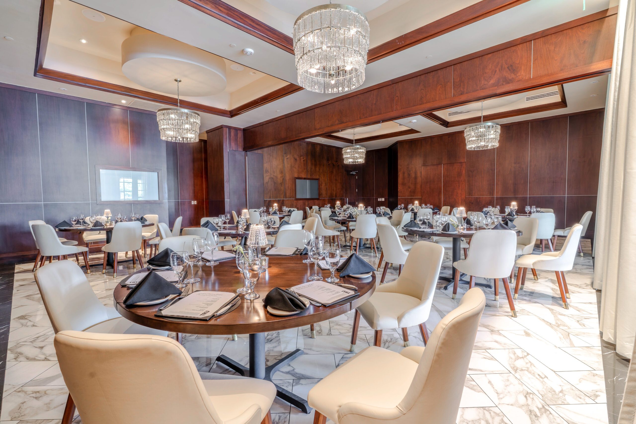 Private dining setup in the Naples Room at Truluck’s The Woodlands, featuring round tables with cream chairs, black napkins, glassware, crystal chandeliers, wood-paneled walls, marble flooring, and polished table settings.