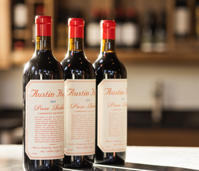Three bottles of Austin Hope Cabernet Sauvignon are displayed on a bar with a softly blurred wine backdrop.