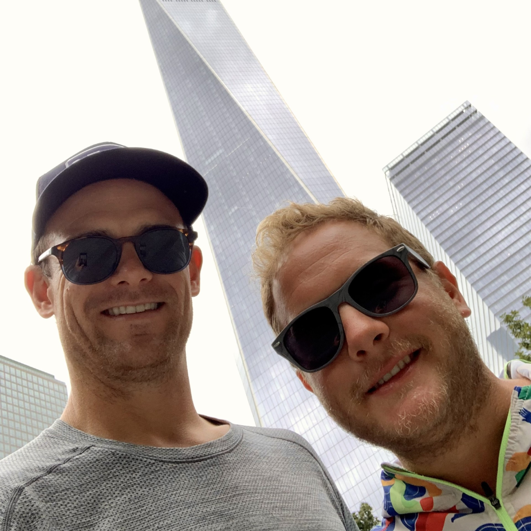 Johnny Griesedieck of Truluck’s Naples posing for a selfie with a friend in front of a tall city skyscraper.