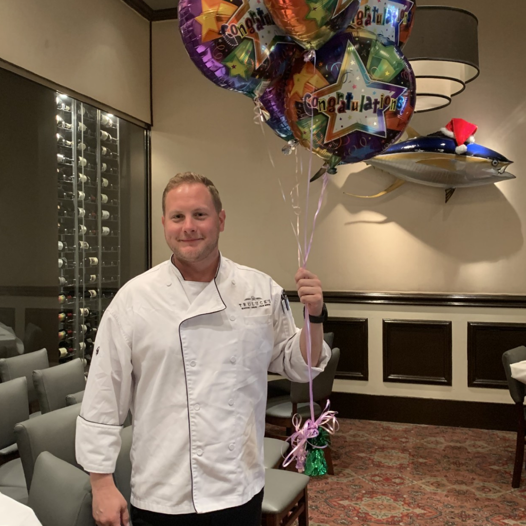 Johnny Griesedieck of Truluck’s Naples wearing a chef coat and holding congratulations balloons inside the restaurant.