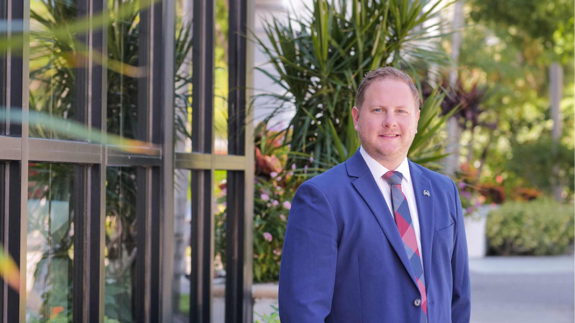 Johnny Griesedieck, General Manager of Truluck’s Naples, standing outdoors in a blue suit and tie near tropical landscaping.