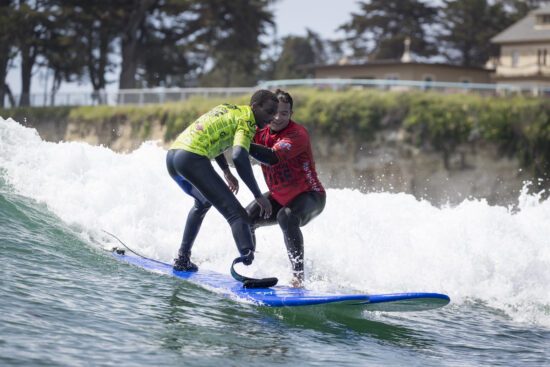 Two surfers ride a wave together during an Operation Surf session, with one participant standing on a blue surfboard while an instructor supports him through the breaking water.