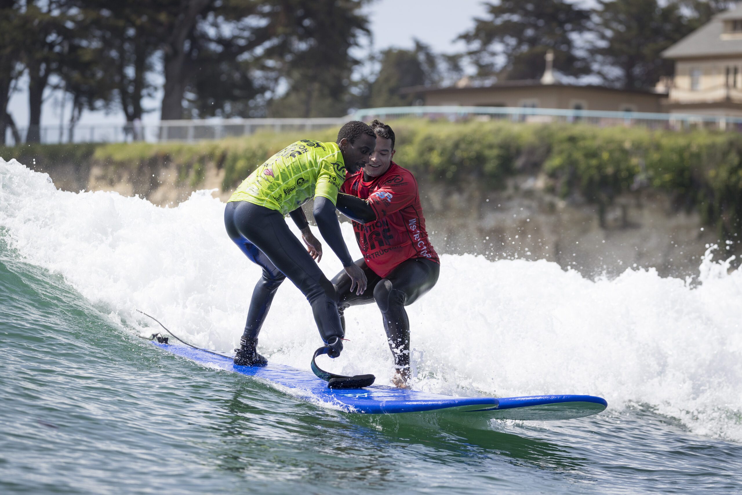 Two surfers ride a wave together during an Operation Surf session, with one participant standing on a blue surfboard while an instructor supports him through the breaking water.