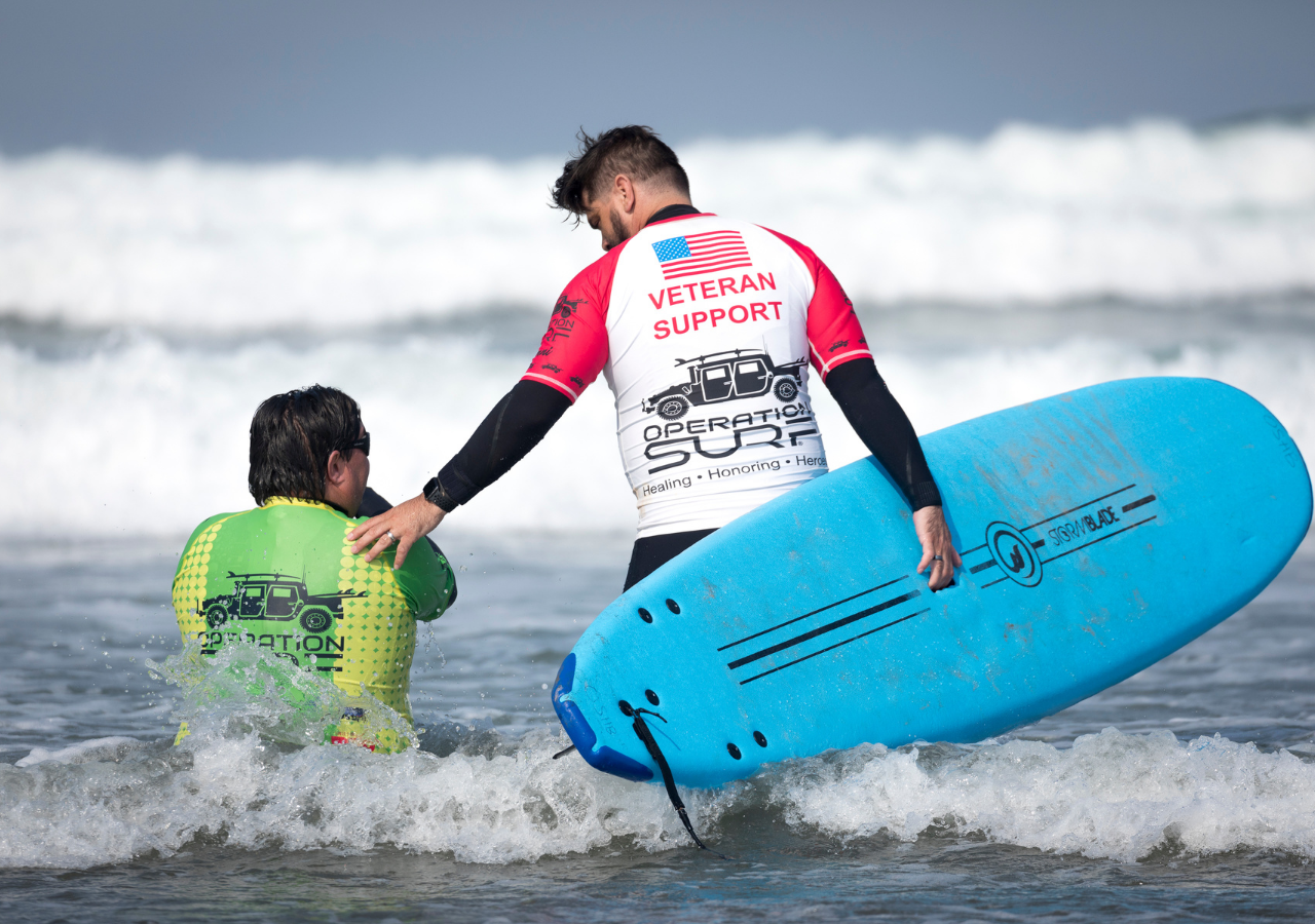 An Operation Surf veteran support volunteer guides a participant into the ocean with a blue surfboard as waves break in the background.