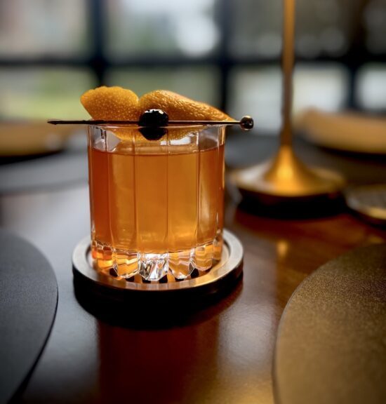 Toasted Pecan Old Fashioned cocktail in a crystal rocks glass, garnished with an orange peel and cherry on a cocktail pick.