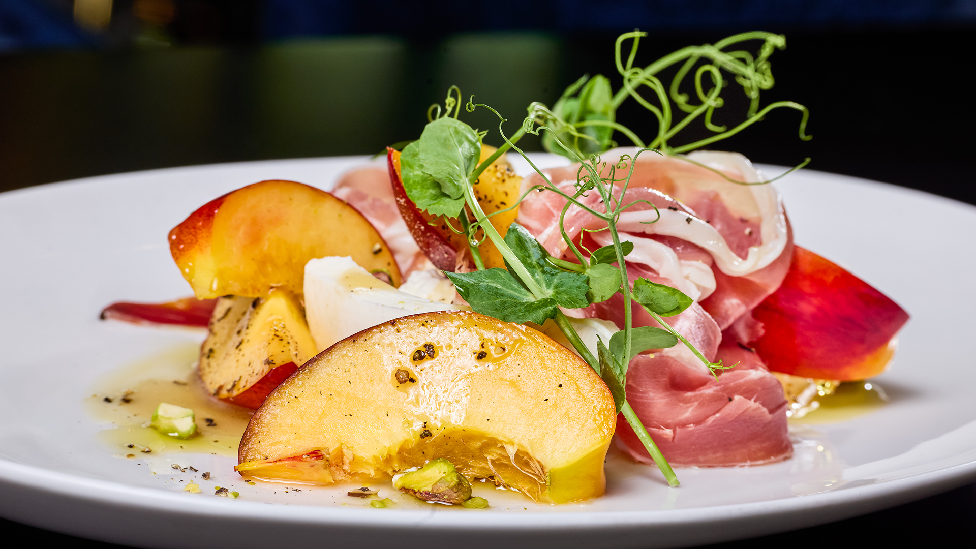 Burrata and stone fruit salad with prosciutto, sliced peaches, pistachios, fresh greens, and olive oil on a white plate.