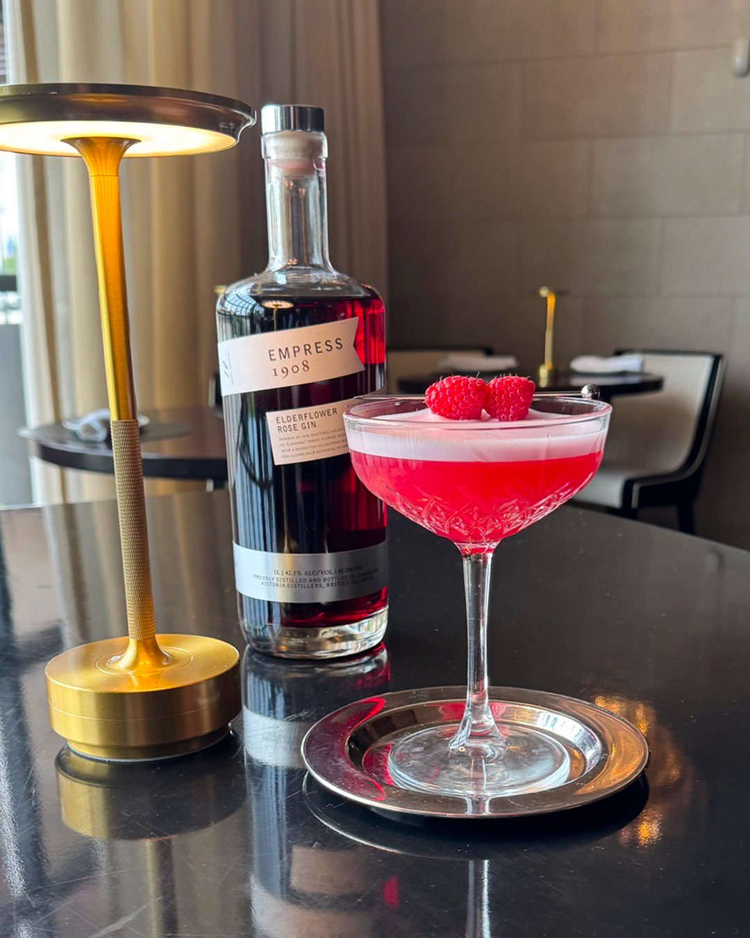 Lychee Empress 1908 cocktail garnished with raspberries beside a bottle of Empress Elderflower Rose Gin on a polished bar table.