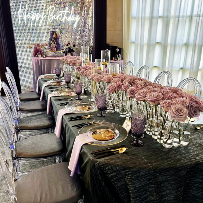 Elegant birthday table setting with blush pink roses, clear chairs, gold-accented place settings, and a glowing “Happy Birthday” sign.